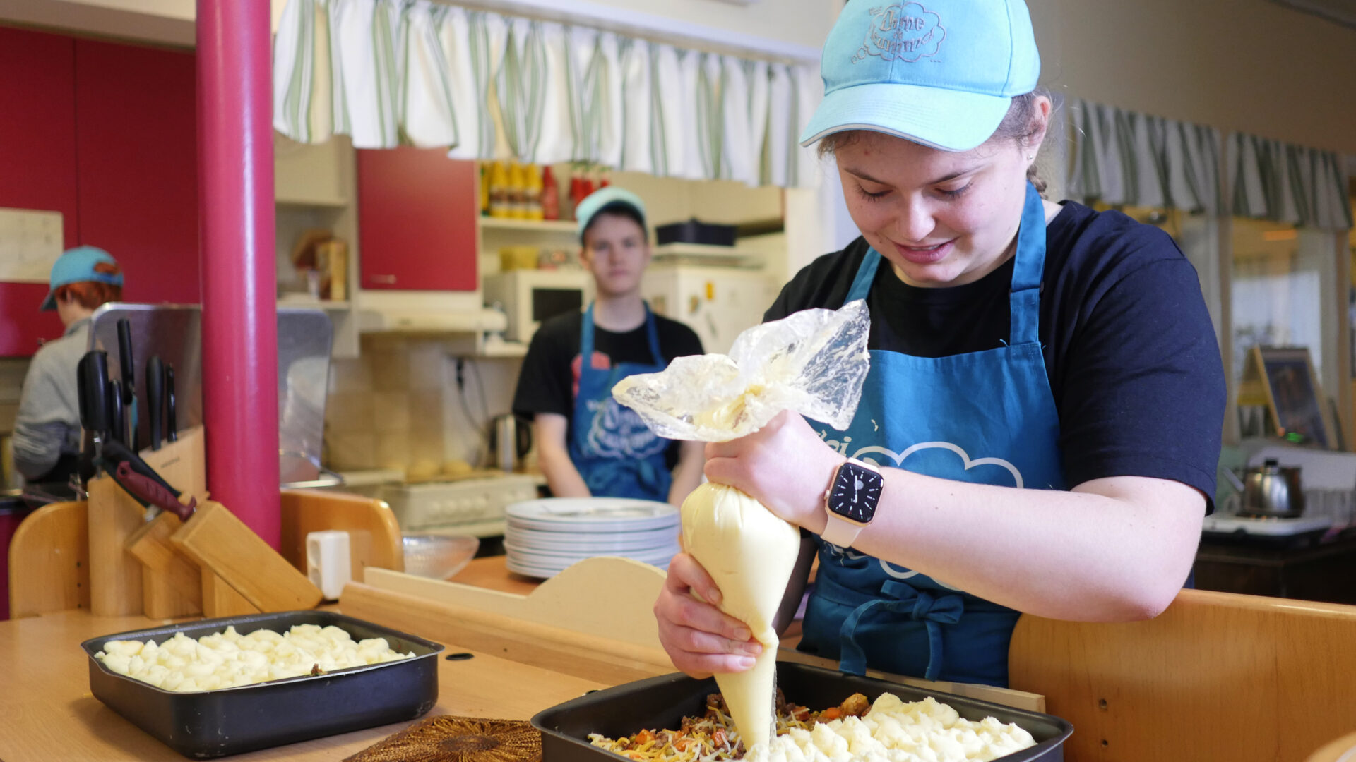 Nuori nainen käyttää pursotinta. Hänellä on päällään sininen essu ja lippis. Nuori mies seuraa tilannetta taustalla.