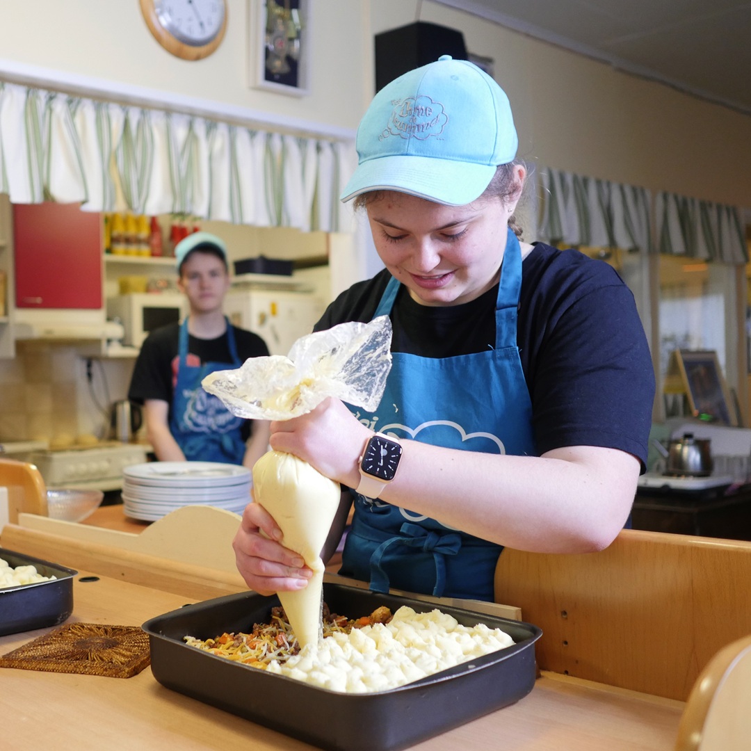 Nuori nainen käyttää pursotinta. Hänellä on päällään sininen essu ja lippis. Nuori mies seuraa tilannetta taustalla.