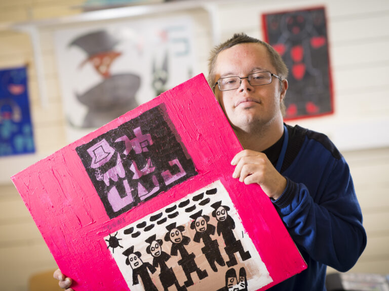 Mies pitää käsissään maalausta, jossa on ihmishahmoja pinkillä taustalla.