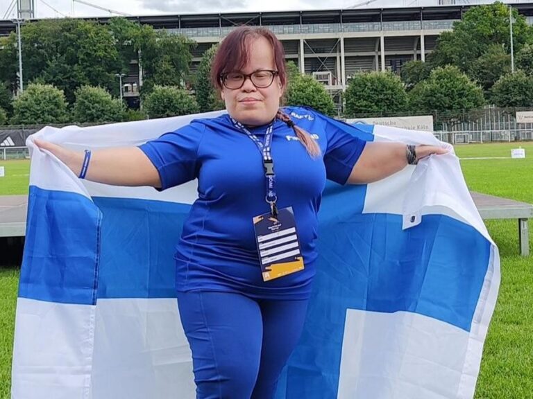 Nainen sinisessä asussa pitää Suomen lippua selkänsä takana.