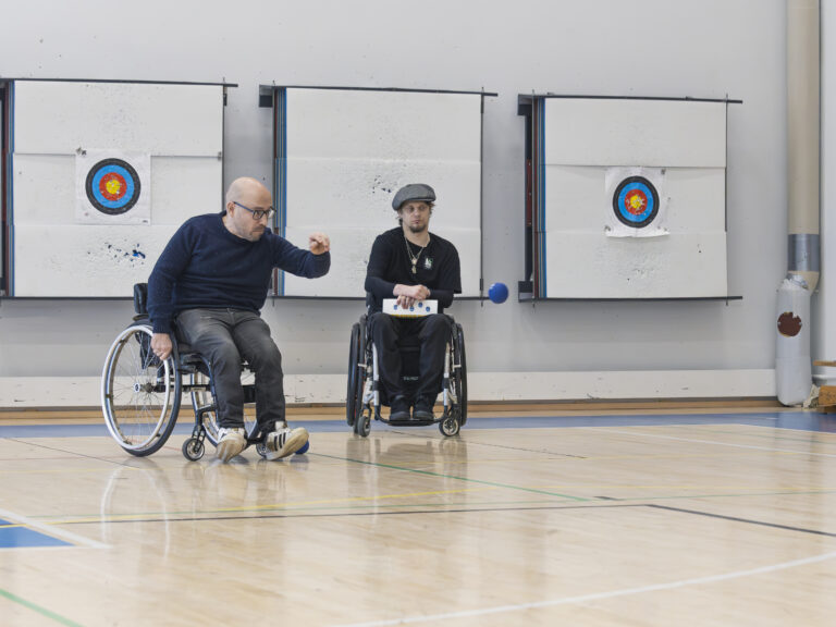 Kaksi miestä pelaa bocciaa pyörätuolista. Mies heittää sinistä palloa.