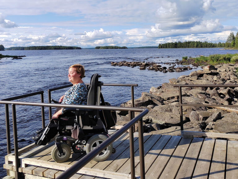 Sanna Kalmari istuu pyörätuolissa laiturilla ja katsoo järvimaisemaa.