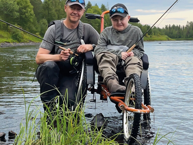 Jere Huovinen istuu kalastukseen sopivassa pyörätuolissa rantavedessä virveli kädessä. Vierellä isä Teemu Huovinen.